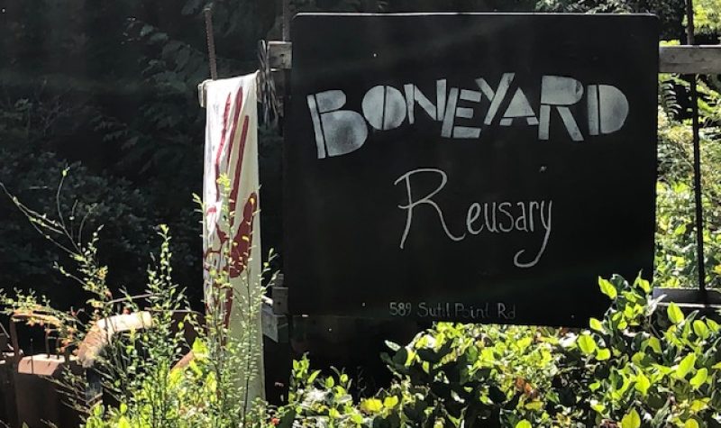 A black sign with white letters in a forested setting marks the entryway to the Boneyard Reusary