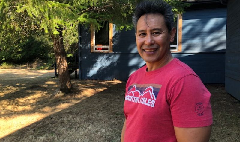 A dark haired man sporting a red t-shirt stands outside a blue building sitting in the shadows of a tree.