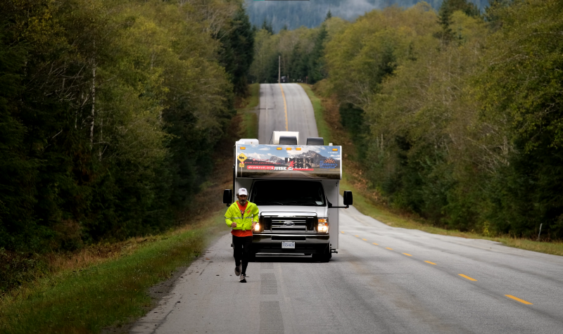 Along a highway at dusk, a man wearing hi-viz running gear is running in front of a 'support vehicle' which is a 35-foot RV follows behind him.