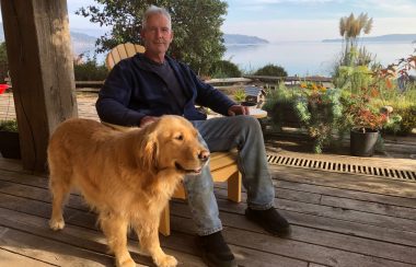 A man and his dog sit on a deck with an ocean view in the background.
