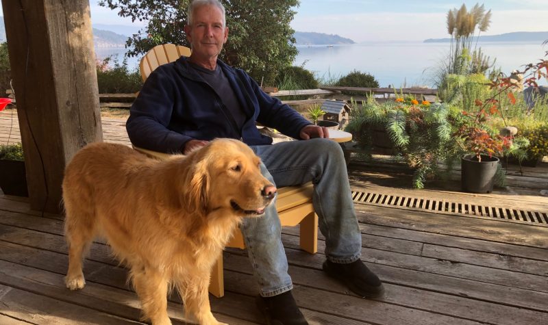 A man and his dog sit on a deck with an ocean view in the background.