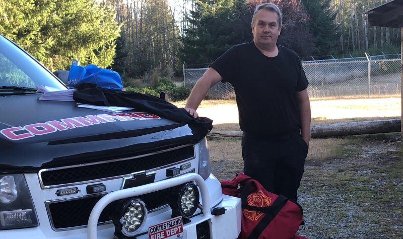 A man dressed in dark clothing leans on a truck with red duffle bags at his feet.