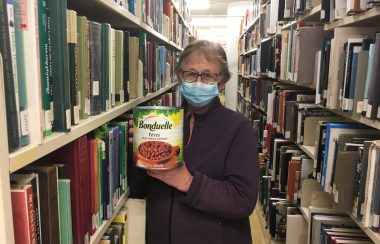 A woman wearing a COVID mask, holding a very large can of beans, standing in between large shelves of books.