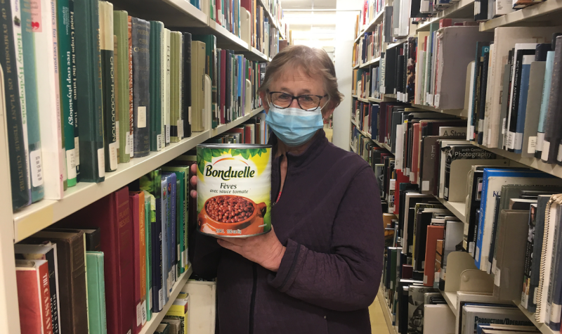 A woman wearing a COVID mask, holding a very large can of beans, standing in between large shelves of books.