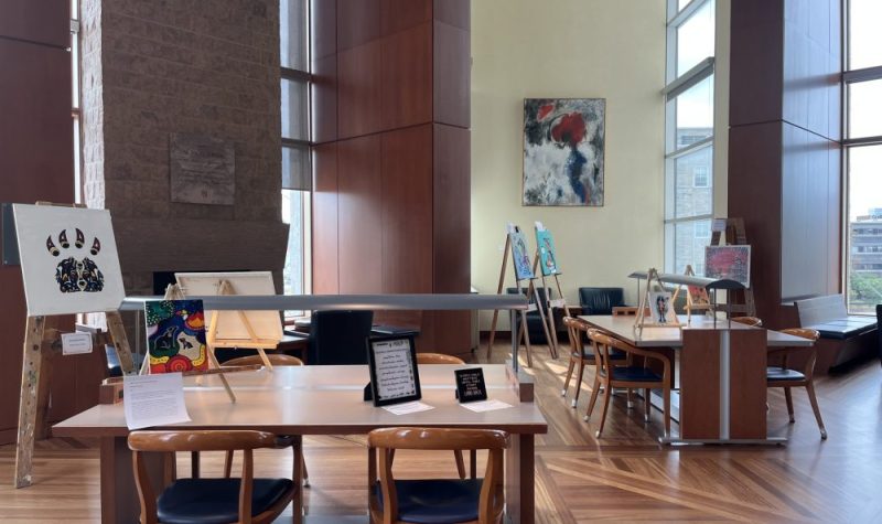 A room with large windows, wooden floors, and a few tables with chairs scattered throughout the room. There are easels with paintings set up throughout the room and a few art peices on the tables.