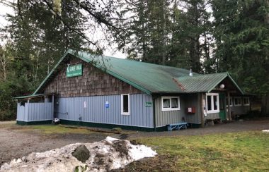 A one story wooden building with a green roof sits in a forested setting.