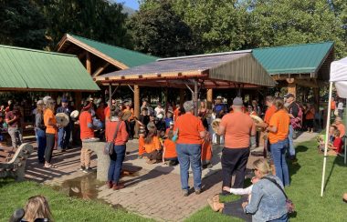 A group of people wearing orange shirts are gathered around a wind shelter in a sunny Lakeside Park as a drum circle is held