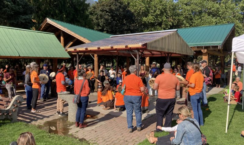 A group of people wearing orange shirts are gathered around a wind shelter in a sunny Lakeside Park as a drum circle is held