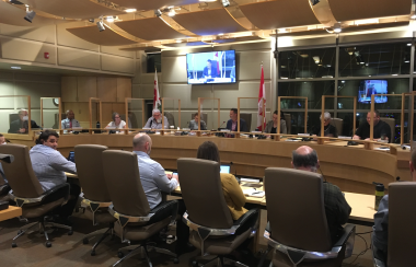 The inside oThe interior of a city council chambers. A group of councillors sit around a table. There are windows in the background and a tv and flags.