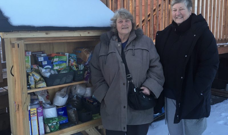 Two women stand beside a cupboard with open doors, filled with various foodstuffs and supplies. It's winter, and the cupboard has a roof with snow on it.