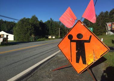 A bright orange construction sign with a stick man holding a sign next to a sunny highway in Nova Scotia