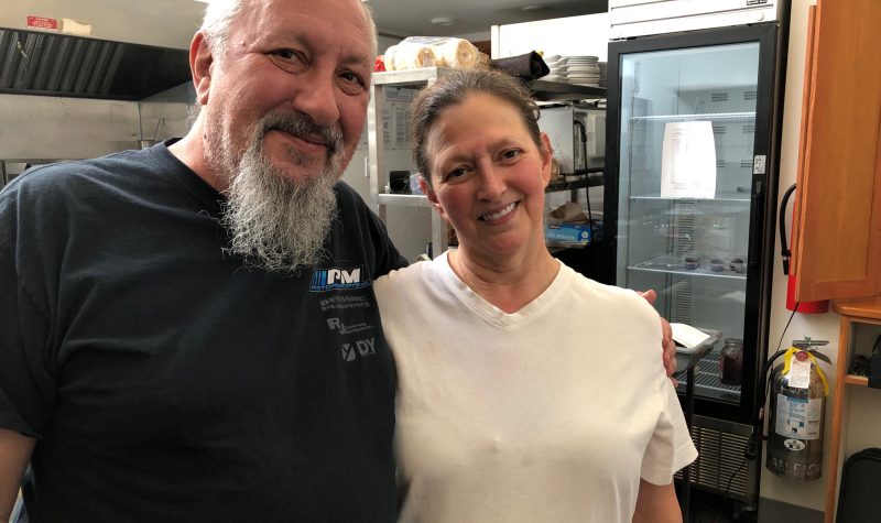 A man with a grey beard wearing a black t-shirt and a woman clad in a white blouse stand in a commercial kitchen.