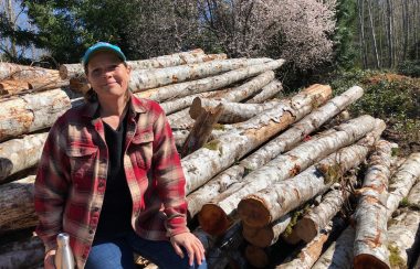 A woman wearing a ball cap and plaid shirt sits atop a pile of logs in a rural setting.