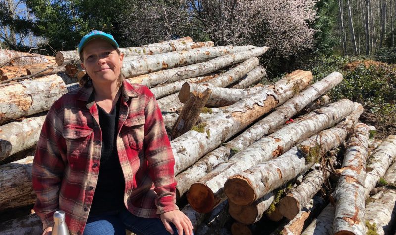 A woman wearing a ball cap and plaid shirt sits atop a pile of logs in a rural setting.