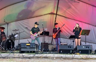 Quatre jeunes sont sur scène. Il y en a un à la batterie, deux à la guitare électrique, et une autre chante. Derrière eux, un arrangement de lumières multicolore.