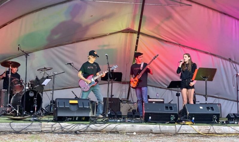 Quatre jeunes sont sur scène. Il y en a un à la batterie, deux à la guitare électrique, et une autre chante. Derrière eux, un arrangement de lumières multicolore.