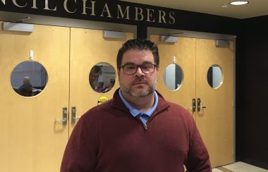 A man standing in front of doors with the words Council Chambers above them.