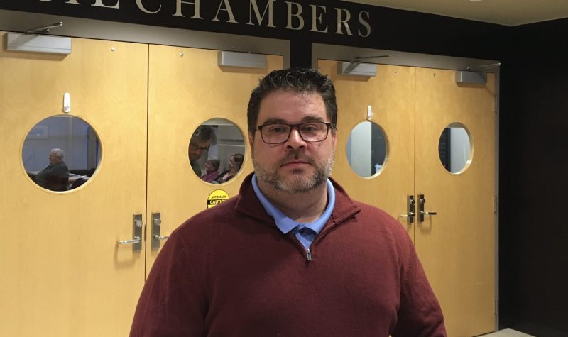 A man standing in front of doors with the words Council Chambers above them.