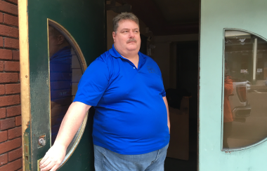 A man in blue shirt stands at green doors with half moon windows.