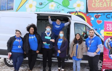 The YEG Youth Connect Team stand in front of their van full of supplies at the iHuman Youth Society. The van is parked in front of a giant mural that's on the iHuman building. Weather was clear.