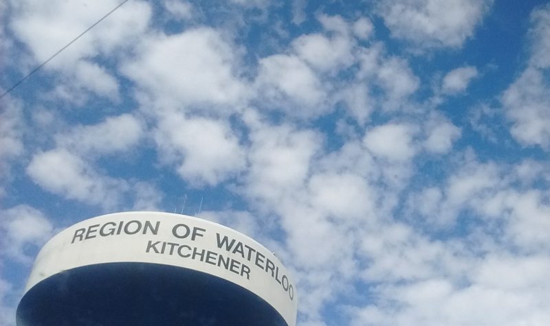 The top of a water tower with the words 'Region of Waterloo Kitchener' against a cloudy blue sky.