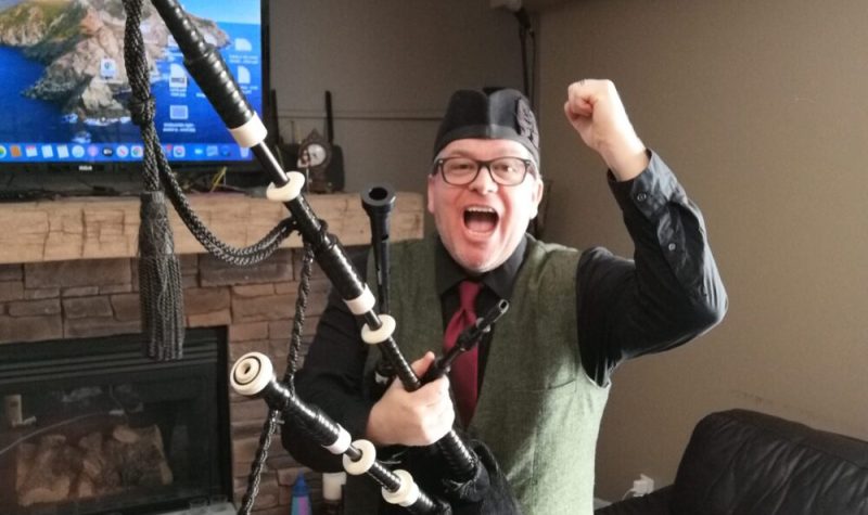 Peter Hummel holds his left fist in the air while holding bagpipes in his right hand and standing in a living room with a television on in the background