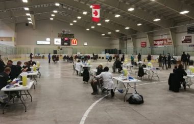 People sit at folding tables inside of the Tantramar Memorial Civic Centre. People in white coats are administering shots.