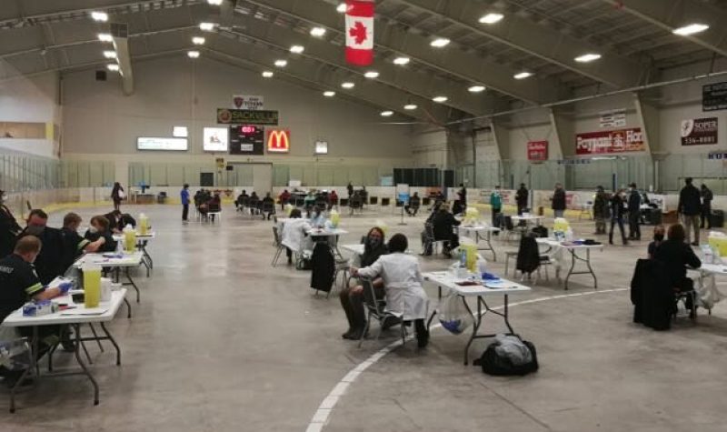 People sit at folding tables inside of the Tantramar Memorial Civic Centre. People in white coats are administering shots.