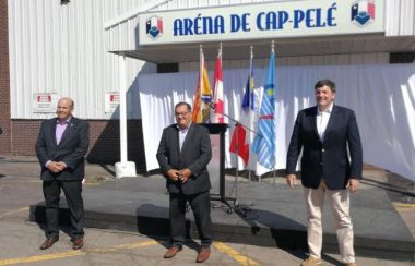 Three men in suits stand in front of the Arena de Cap-Pelé, with flags in the background.