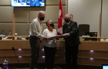 An older couple receive a gift from a man in a suit, all wearing COVID masks.