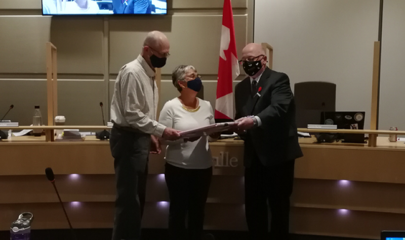 An older couple receive a gift from a man in a suit, all wearing COVID masks.