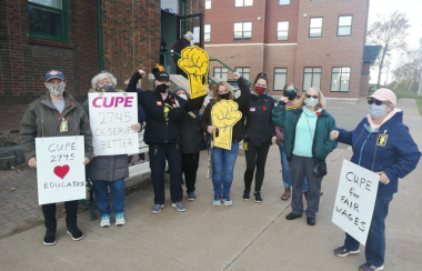 A group of people hold protest signs at a CUPE rally in downtown Sackville on a sunny day.