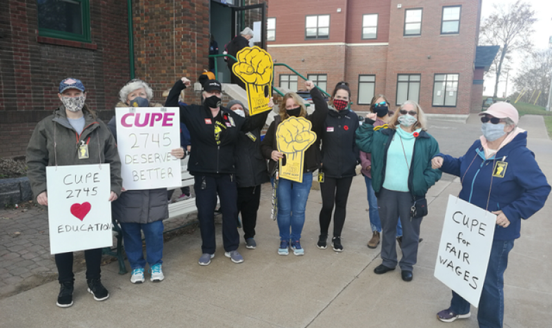 A group of people hold protest signs at a CUPE rally in downtown Sackville on a sunny day.