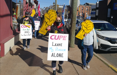 A group of protesters walk down the street in Sackville on a sunny day. They hold protest signs about CUPE.