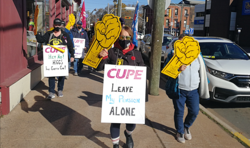 A group of protesters walk down the street in Sackville on a sunny day. They hold protest signs about CUPE.