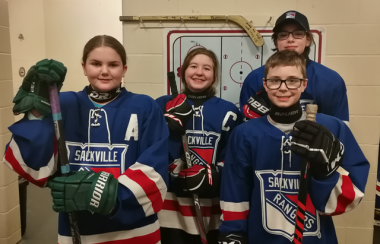 A group of young hockey players stands in an arena hallway. They are wearing blue jerseys with white and red trim.