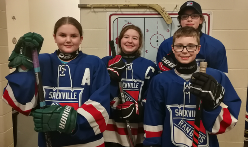 A group of young hockey players stands in an arena hallway. They are wearing blue jerseys with white and red trim.