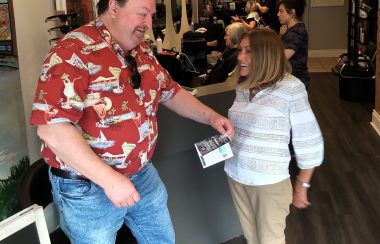 A man in a red shirt and blue jeans gives a flyer to a persn wearing a white and grey shirt and beige pants.