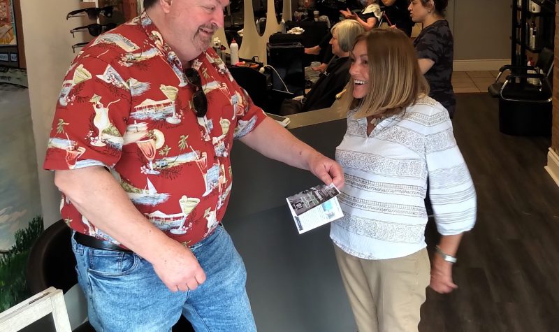 A man in a red shirt and blue jeans gives a flyer to a persn wearing a white and grey shirt and beige pants.