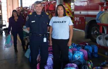 Le chef des pompiers de Lac La Biche, JOhn Kokotilo, et l'agente de soutien aux étudiants du Friendship Centre de Lac La Biche, Tashina Paul, sourient pour la caméra devant des sacs d'école remplis de fournitures scolaires, pendant que des bénévoles en remplissent davantage en arrière-plan.