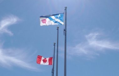 Le drapeau canadien et le drapeau franco-albertain flottent au vent, sur fond de ciel bleu avec quelques nuages.