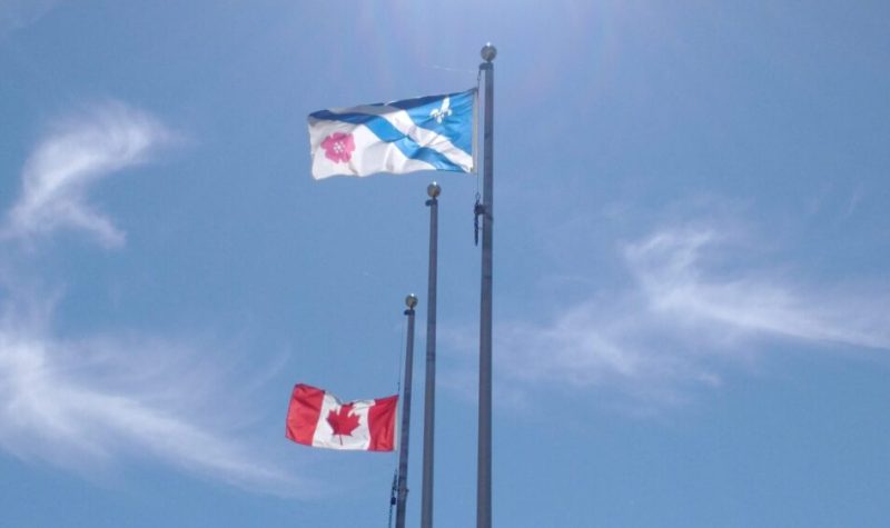 Le drapeau canadien et le drapeau franco-albertain flottent au vent, sur fond de ciel bleu avec quelques nuages.