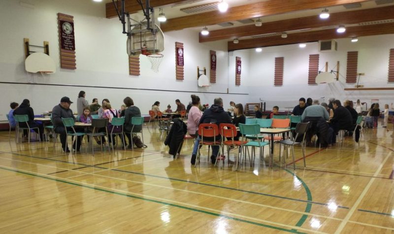 Plan large sur le gymnase de l'École Sainte-Catherine, avec des tables au premier plan et des enfants qui jouent à l'arrière-plan.