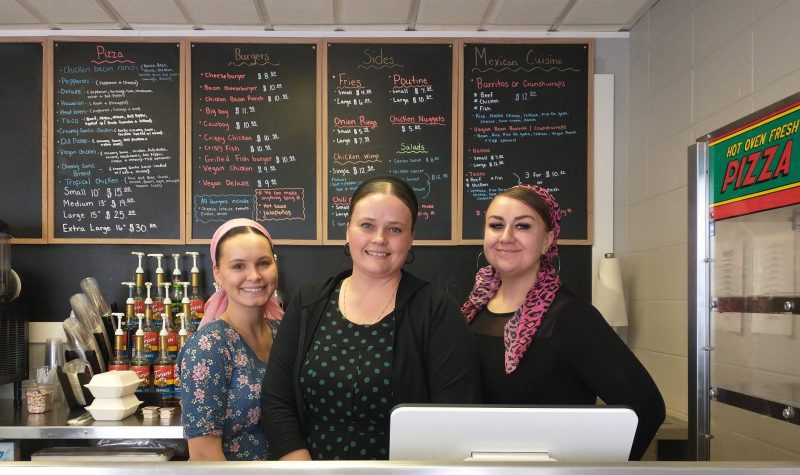 Trois femmes sourissent derrière un comptoir de service, le menu du restaurant en arrière-plan.