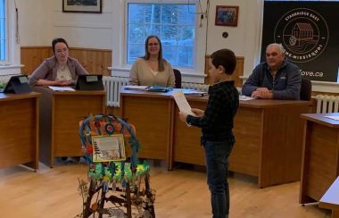 A young boy delivering a chair to the council members. They are sitting behind tables while the boy reads off a paper.