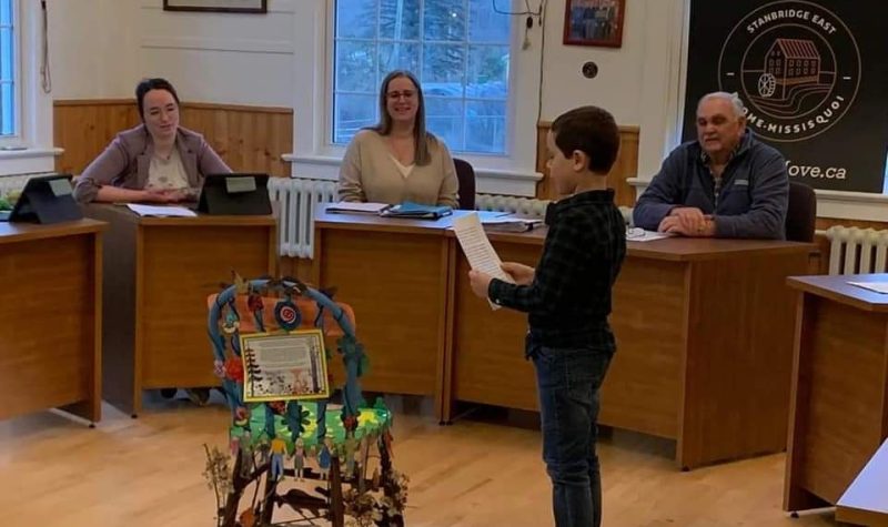 A young boy delivering a chair to the council members. They are sitting behind tables while the boy reads off a paper.