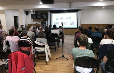 Des gens assis dans une salle devant un projecteur d'information et un micro pour poser des questions en plen milieu de la salle.