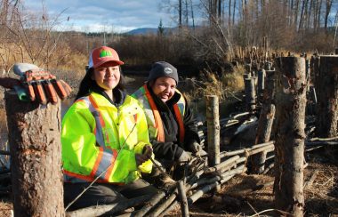 two young people are standing by a fence made of branches near a river