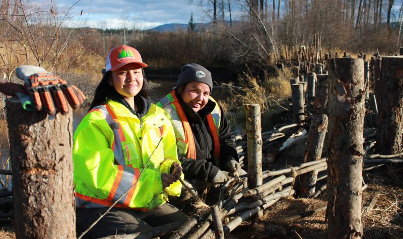 two young people are standing by a fence made of branches near a river