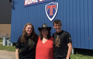 A teacher in a cowboy hat, flanked by two student, all smiling, standing in front of a building with TRHS Titans logo.